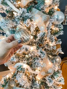 a hand holding a glass ornament in front of a christmas tree with white and silver ornaments