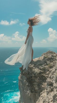 a woman standing on top of a cliff next to the ocean with her hair blowing in the wind