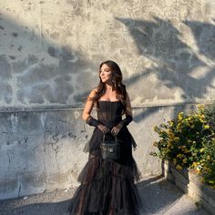 a woman standing in front of a wall wearing a black dress and holding a purse