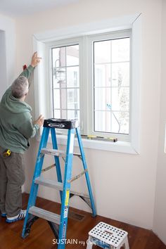 a man standing on a ladder in front of a window while painting the wall with white paint