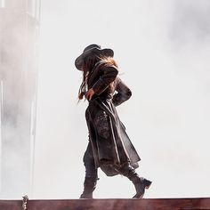 a man with long hair wearing a hat and leather coat walking across a wooden plank