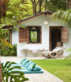 a small white house with wooden shutters on the front and side windows, surrounded by lush greenery