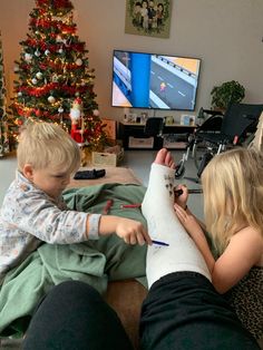 two children are sitting on the couch playing with an arm cast in front of a christmas tree