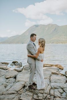 a bride and groom standing on rocks by the water looking at each other with mountains in the background