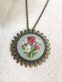 a necklace with flowers on it is hanging from a metal ball chain and sits on top of a white cloth