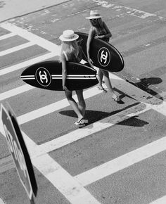 two women walking across a street holding surfboards in their hands and one carrying a hat on her head