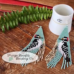 the beaded earrings are next to a cup and fern leaf on a wooden table