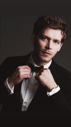 a man in a tuxedo adjusts his bow tie while looking at the camera