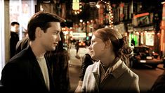 a man and woman standing in the middle of a city street at night, talking to each other