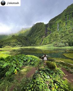 a woman standing on the side of a lush green hillside next to a lake and waterfall