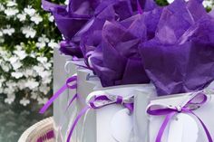 two white bags with purple ribbons and tags on them are sitting in front of some flowers