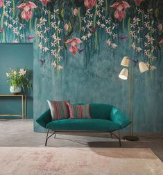 a green couch sitting in front of a wall with pink flowers on it and a lamp next to it