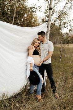 a pregnant couple cuddles against a white sheet in a field with tall grass