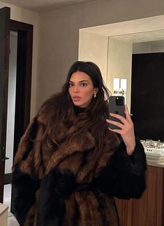 a woman taking a selfie in a bathroom mirror wearing a fur coat and holding a cell phone