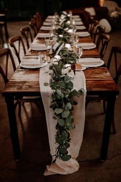 the long table is set with white plates and place settings, along with greenery