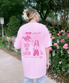 a girl in pink shirt standing on sidewalk next to flowers and trees with her back to the camera