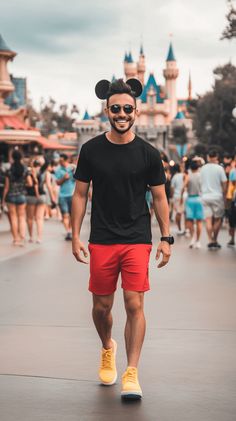 A man wearing a black shirt, red shorts, and yellow shoes, with Mickey Mouse ears. He is smiling and walking through a lively Disney park. Disney outfit. Men’s Mickey Mouse Costume Diy, Mens Mickey Mouse Shirt, Mickey Mouse Outfit Men, Disney Outfits For Guys, Male Disney Outfits, Men Disneyland Outfit, Disney Bounding For Men, Disney Ears For Men, Mens Disney Outfit Ideas