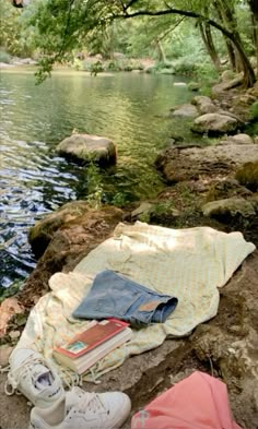 a blanket, book and shoes are sitting on the rocks next to a river