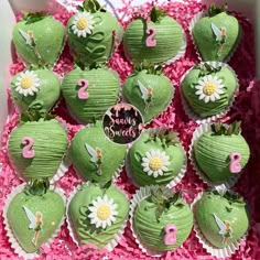 there are many green cupcakes in the box with pink flowers on them and decorations