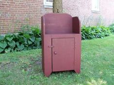 a red cabinet sitting in the grass next to a tree and brick building with windows