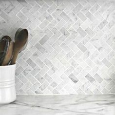 a white vase filled with silver spoons on top of a counter next to a marble tiled wall