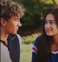 a young man and woman sitting next to each other in front of some trees on the grass