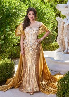 a woman in a gold dress standing next to some bushes and trees with her hands on her hips