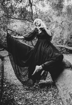 a woman in a long black dress is sitting on a fallen tree branch and posing for the camera