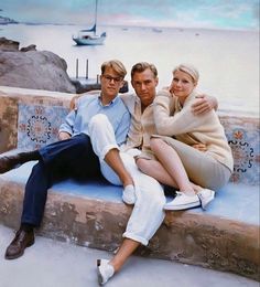 two people are sitting on a bench near the water with boats in the background and one person is holding his arm around the woman's neck