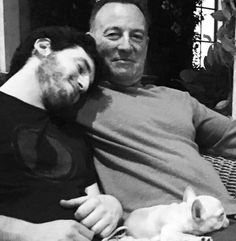 two men sitting on a couch cuddling with a small white dog in front of them