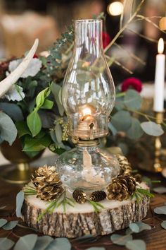 a table topped with a glass vase filled with pine cones and greenery next to a candle