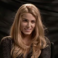 a woman with long blonde hair sitting in front of a black backdrop smiling at the camera