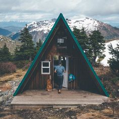 two people are standing in the doorway of a small cabin with mountains in the background