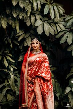 a woman wearing a red and white sari standing in front of some plants with her hands on her hips