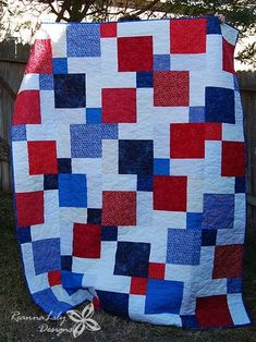 a red, white and blue quilt sitting on top of a grass covered field next to a fence