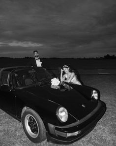 a man and woman sitting on top of a black sports car in the middle of a field