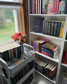 a book shelf filled with lots of books next to a window and a vase full of flowers