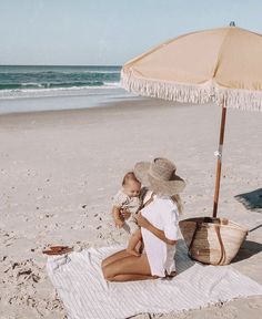 a woman sitting on top of a beach next to a baby under an open umbrella