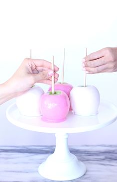 two hands are holding apples on a white cake plate with toothpicks in them