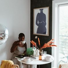 a woman sitting at a white table drinking from a cup and holding a coffee mug