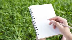 a person holding a pen and writing on a notebook in front of some green grass