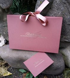 a pink wedding card with a bow on the front and side, sitting next to a rock