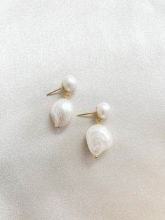 two white pearls are hanging from gold ear pins on a white surface, with one pearl in the foreground