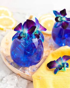 two blue vases filled with purple flowers sitting on top of a wooden tray next to lemon slices