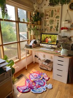 a desk with a computer on top of it in front of two windows and plants