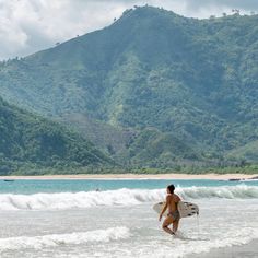 a woman walking into the ocean with a surfboard in her hand and mountains in the background