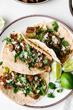 three tacos on a plate with limes, cilantro and other ingredients