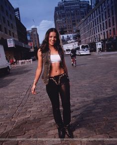 a woman in black pants and white top walking down a brick street with tall buildings behind her