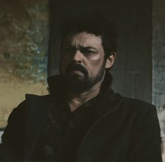 a man with a beard and black jacket looking at the camera while standing in front of a wall