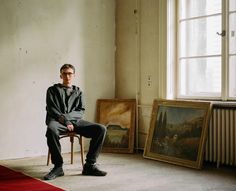 a man sitting on top of a chair in front of a window next to paintings